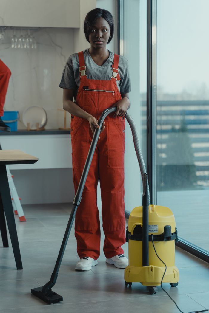 services-04 An adult woman in red coveralls operates a vacuum cleaner in a modern indoor setting, focusing on cleaning tasks.