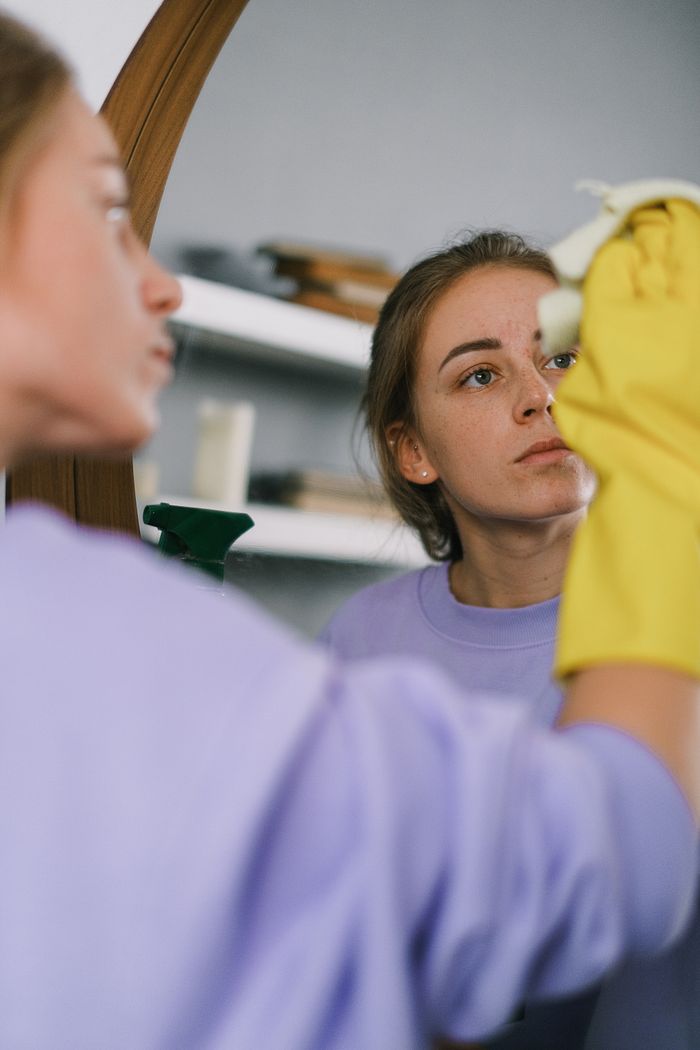 heros-img Crop female in purple sweatshirt and latex gloves washing mirror in light apartment in daytime